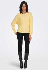 Geel geribd truitje met een brede halslijn en doorlopende schouders, gecombineerd met zwarte skinny jeans en kniehoge zwarte leren laarzen.