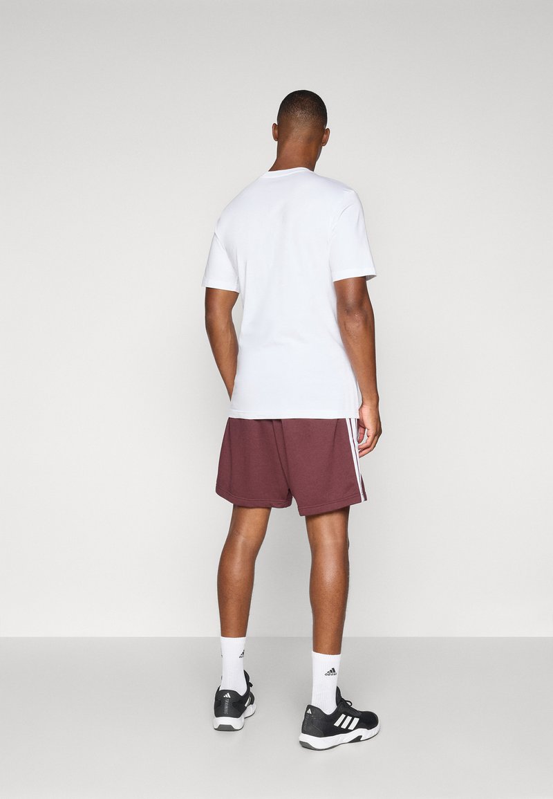 Camiseta blanca de manga corta y pantalones cortos granate con rayas blancas en los laterales, combinados con zapatillas deportivas negras y calcetines blancos.