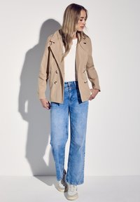 Beige dubbelzijdig trenchcoat over een wit shirt, gecombineerd met lichte spijkerbroeken met wijde pijpen en grijze sneakers, tegen een neutrale achtergrond.