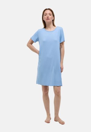Femme debout pieds nus portant une robe bleu clair au genou avec des manches courtes sur un fond blanc uni.