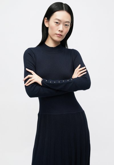 Sukienka dzianinowa w kolorze granatowym, z dopasowanym górnym partem, plisowaną spódnicą oraz długimi rękawami ze zdobieniami w postaci guzików na mankietach. Gładka tekstura na całej powierzchni.