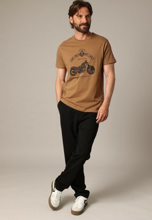 Bruine katoenen t-shirt met zwarte motorfietsafbeelding en tekst. Gecombineerd met zwarte broek en witte sneakers met bruine accenten.