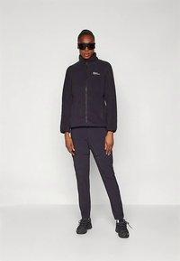 Czarna kurtka polarowa z frontowym zamkiem błyskawicznym, stójką i logo. Stylizowana z pasującymi ciemnymi spodniami oraz czarnymi butami sportowymi.