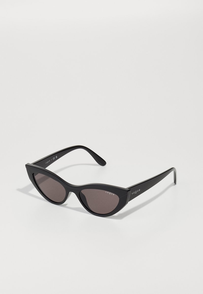 Ochelari de soare cu formă de cat-eye, cu o ramă neagră mată și lentile închise la culoare, având logo-ul "vogue" pe lateral și o formă unghiulară distinctivă.