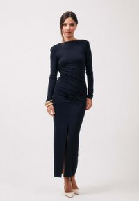 Femme debout portant une robe noire moulante à manches longues avec des détails froncés, une fente avant, des talons blancs et des bracelets empilés sur son poignet droit.