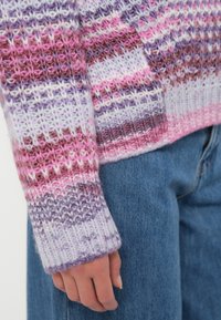 Hand resting by side, wearing a multicolored knit sweater in pink, purple, and white stripes paired with blue denim jeans.