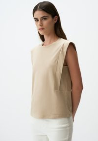 Beżowy top bez rękawów wykonany z miękkiego materiału, z okrągłym dekoltem, posiadający strukturalne akcenty na ramionach oraz boczną kieszeń.