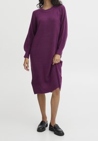 Abito a maglia a maniche lunghe di un profondo viola, con texture a coste e spacco laterale. Abbinato a mocassini neri lucidi.
