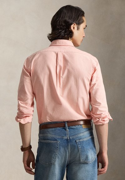 Vīrietis ar tumšiem matiem, valkājot sarullētu rozā garroku kreklu, zilas džinsas, brūnu jostu un pulksteni, stāvot pret neitrālu fonu.