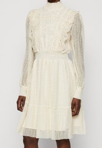 Krämfärgad klänning med spetsdetaljer, volangkrage, långa genomskinliga ärmar och prickig textur. Snyggt format midja med elastisk, lager-på-lager kjol.