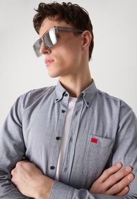 Cămașă gri cu nasturi, dotată cu un buzunar aplicat și un logo roșu, purtată peste un tricou alb. Ochelari de soare transparenți cu lentile reflective.