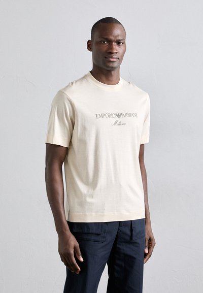 Beige pamut póló rövid ujjakkal, szürke színben "EMPORIO ARMANI Milano" felirattal. Klasszikus kerek nyakú kialakítás.