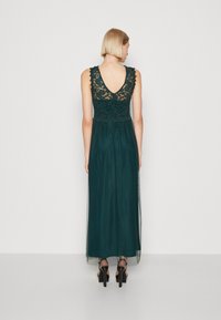 Rochie lungă de culoare verde închis, cu un corp din dantelă, spate în formă de V și fustă fluidă. Prezintă detalii din dantelă și pantofi cu toc înalt de culoare neagră.