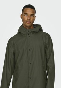 Jachetă impermeabilă de culoare verde măslin, cu glugă, butoane frontale și buzunare laterale. Textură netedă și design minimalist.