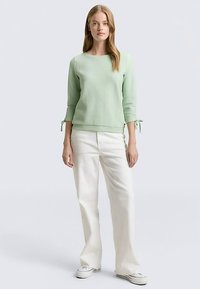 Hellgrünes, strukturiertes Strickoberteil mit dreiviertel Ärmeln und Bindebändern an den Handgelenken, kombiniert mit weißen, weit geschnittenen Hosen und lässigen Schuhen.