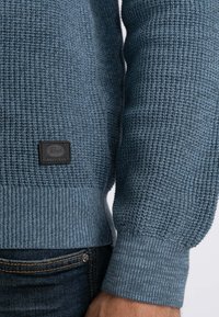 Blauwe gebreide trui met een gestructureerd patroon, ribbels aan de mouwen en een zwart label aan de onderrand. De stof toont subtiele kleurnuances.