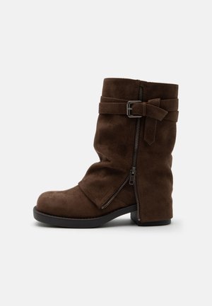 Botte mi-mollet en daim marron avec fermeture éclair latérale, petit talon bloc et bride décorative avec boucle métallique sur fond blanc.