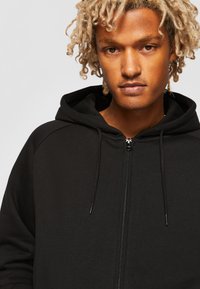 Sort zip-hoodie med lynlås foran, snore i hætten og tekstureret stof. Har raglanærmer og et enkelt design.
