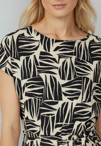Kleid mit einem cremefarbenen Hintergrund und einem schwarzen Blättermuster, kurzen Ärmeln und einem runden Ausschnitt. Der Stoff hat eine strukturierte, zerknitterte Optik.