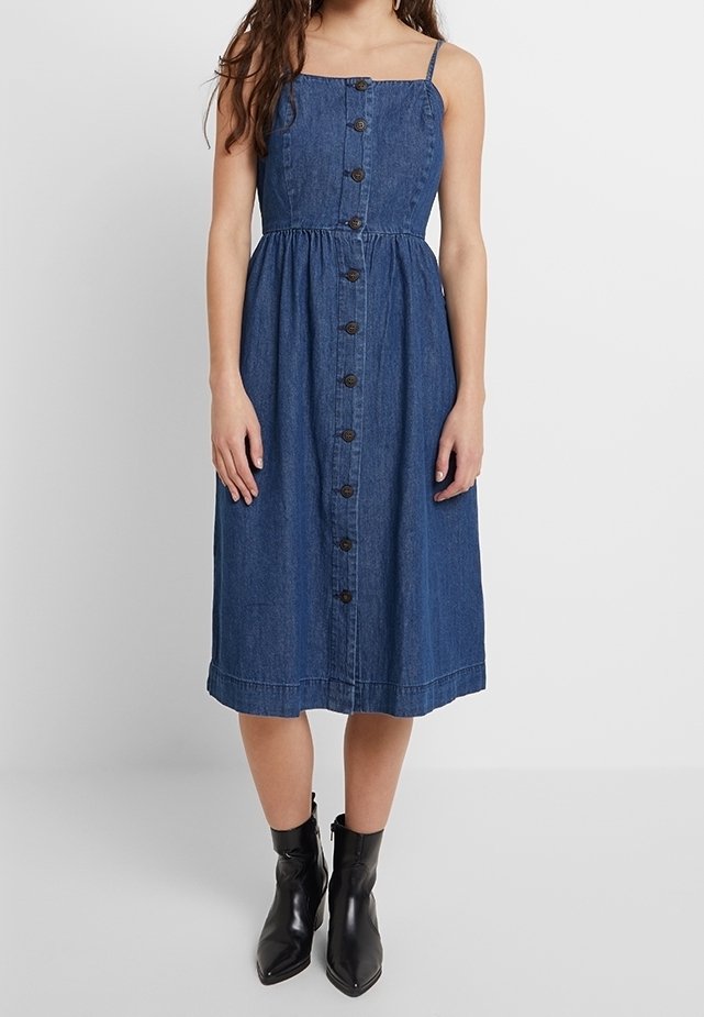 Femme portant une robe midi en denim bleu sans manches avec des boutons sur le devant, associée à des bottes noires à la cheville, se tenant devant un fond blanc uni.