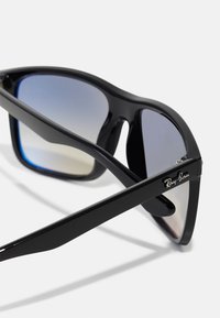 Ochelari de soare din plastic negru cu lentile degrade cu nuanțe de albastru. Formă rectangulară cu partea superioară curbată. Logo-ul Ray-Ban pe braț.