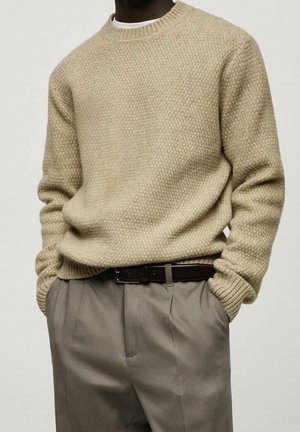 Mand iført en tekstureret beige strikket sweater over en hvid skjorte, stoppet ned i beige plisserede bukser med et mørkebrunt bælte, hænderne i lommerne.