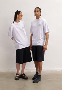 Two individuals posed with white oversized t-shirts featuring a sun graphic and black shorts. Footwear includes black sandals and sneakers.