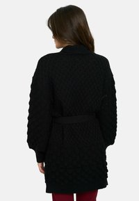 Cárdigan negro de punto con patrón texturizado en relieve, mangas largas y cintura ceñida con cinturón. Presenta un cuello ancho y bolsillos.