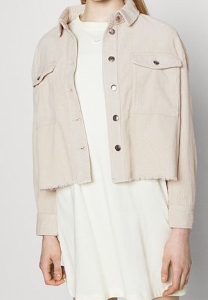 Personne portant une veste courte en velours côtelé beige avec des boutons, par-dessus une robe blanche ornée d'un petit logo Nike près du col.