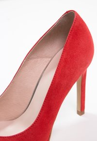 Chaussure à talon haut pour femme en daim rouge, avec un bout pointu et une doublure intérieure beige, présentée sur un fond blanc.