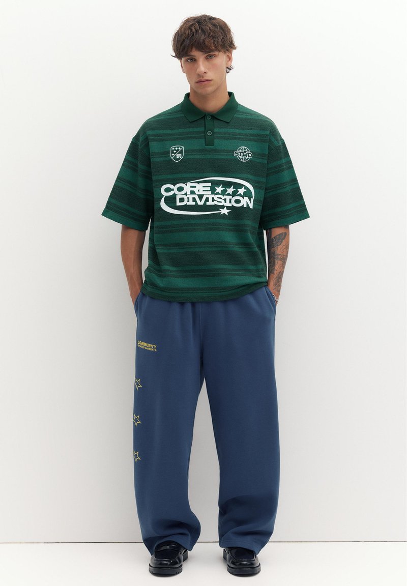 Jeune homme aux cheveux courts et bouclés, portant un polo vert rayé avec des logos et un pantalon bleu ample avec des motifs d'étoiles, debout sur un fond blanc.