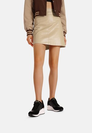 Sneaker in bianco e nero con suola spessa, abbinato a una mini gonna in ecopelle beige e a una giacca bomber marrone e beige.