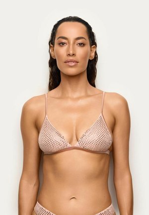 Mujer con cabello largo y oscuro que lleva un top de bikini triangular de malla beige y braguitas a juego contra un fondo claro liso.