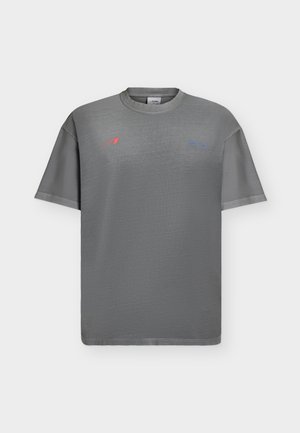 T-shirt grigia a maniche corte con scollo rotondo e piccoli loghi rossi e blu sulla parte superiore del petto su sfondo semplice.