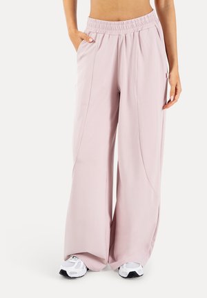 Kobieta nosząca luźne, jasnoróżowe spodnie z szerokimi nogawkami, elastycznym pasem i białe tenisówki, stojąca z jedną ręką w kieszeni.