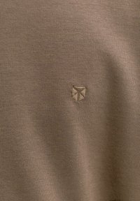 Κοντινή λήψη καφέ υφάσματος με μικρό κεντημένο λογότυπο σε σχήμα ασπίδας ραμμένο με νήμα στο ίδιο χρώμα κοντά στο κέντρο.