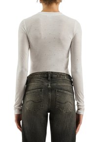 Langärmeliges, geripptes weißes Oberteil mit verzierten silbernen Akzenten. Kombiniert mit dunkelgrauen, ausgewaschenen Denim-Jeans mit Gesäßtaschen und Ziernähten.