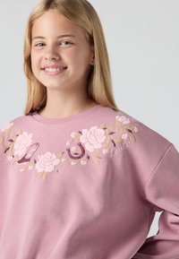 Felpa rosa chiaro con ricami floreali e dettagli giocosi, presenta un collo a giro e un tessuto morbido e texturizzato. Progettata per il comfort.