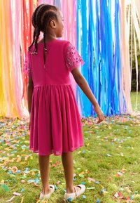 Roze jurk met korte, doorschijnende, geborduurde mouwen, een getailleerde taille en een uitlopende rok. Draag met glinsterende schoenen. Kleurrijke confetti op de grond.