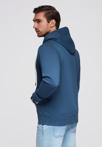 Niebieska bluza z kapturem, z regulowanym sznurkiem, o miękkiej fakturze i z przednią kieszenią typu kangur, w zestawieniu z jasnoniebieskimi dżinsami, widok z tyłu.