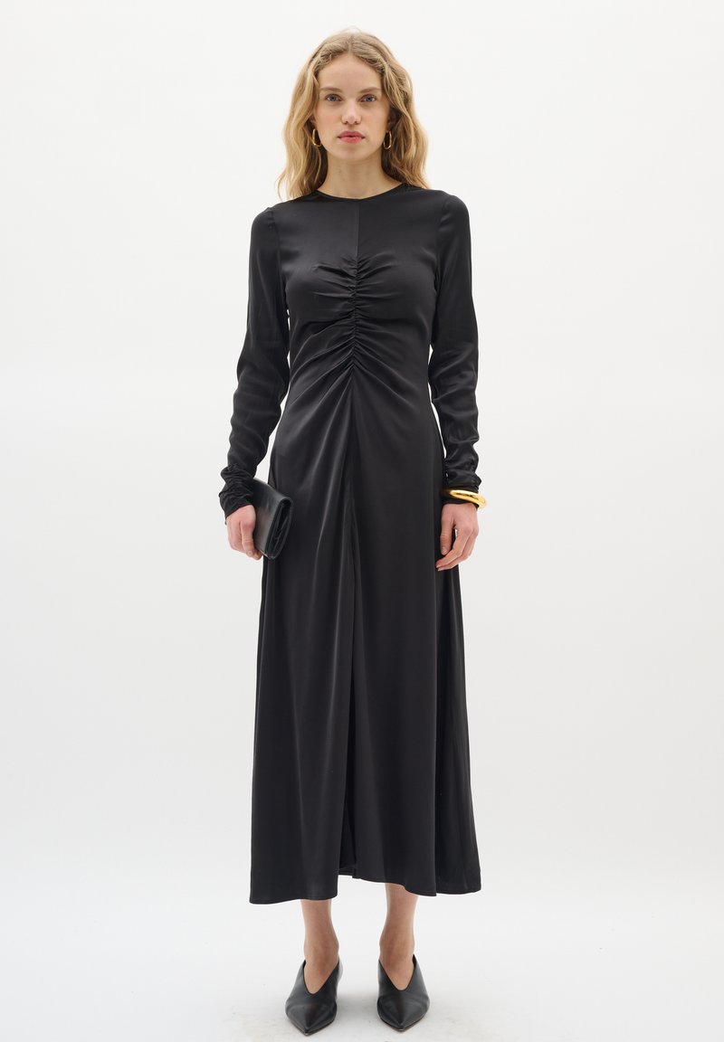 Schwarzes, langärmliges Kleid mit Raffung an der Vorderseite, aus einem glatten Stoff. Kombiniert mit schwarzen spitzen High Heels und einer Clutch.