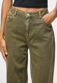 Olīvzaļas bikses, izgatavotas no mīksta denima ar brīvu piegriezumu, piedāvā priekšējās kabatas, metāla pogu un viegli saskatāmus šūšanas elementus.