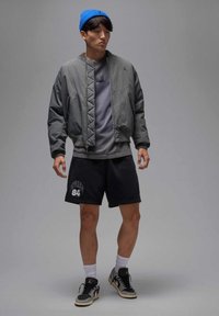 Grijze bomberjas met diagonale stiksels, grijs t-shirt, zwarte korte broek met "Jordan 84," blauwe muts en zwarte sneakers met grijze accenten.