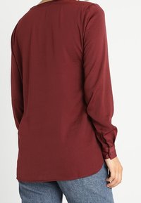 Blusa bordeaux a maniche lunghe con una texture liscia, scollatura rotonda e polsini con bottone, caratterizzata da un leggero orlo asimmetrico.
