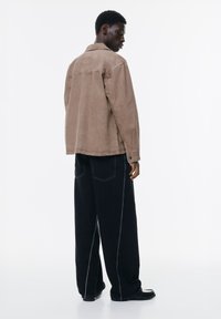 Ruskea corduroy-takki rentoa istuvuutta, jossa on point collar, sivutaskut ja mustat leveälahkeiset farkut, joissa on kontrastivalkoiset tikkaukset.
