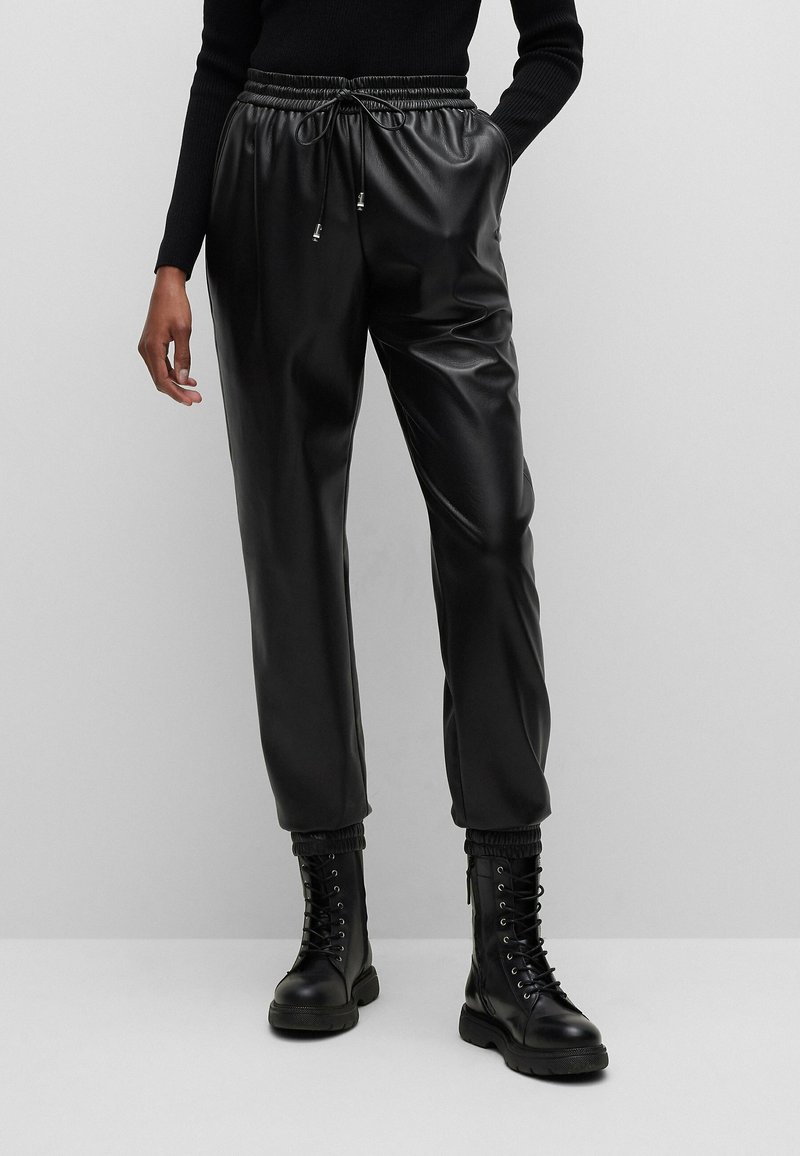 Personne portant un pantalon jogger noir en simili cuir avec des poignets élastiques, un haut noir à manches longues et des bottes de combat noires lacées.