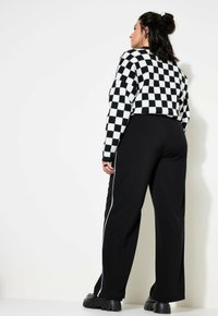 Černobíle károvaný svetr s dlouhými rukávy v kombinaci s černými kalhotami se bílými bočními pruhy. Model stojí otočený zády, aby předvedl outfit.
