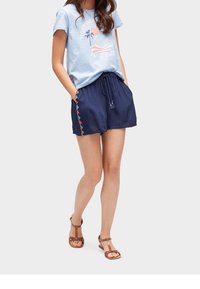 Kvinna som bär en ljusblå grafisk t-shirt, marinblå shorts med snörning och sidomönster, samt bruna sandaler, står med händerna i fickorna.