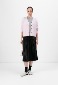 Cárdigan de punto rosa claro con botones, usado sobre una blusa gris y una falda negra con pliegues. Combinado con zapatos blancos sin cordones y calcetines negros.