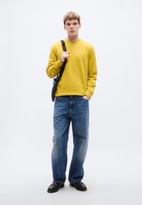 Κίτρινο φούτερ με στρογγυλή λαιμόκοψη, συνδυασμένο με μπλε τζιν παντελόνι. Το μοντέλο φοράει μαύρη τσάντα crossbody και μαύρα παπούτσια.